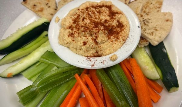 A bowl of hummus dusted with paprika, ringed by warm pita wedges and cucumber, celery and carrot sticks.